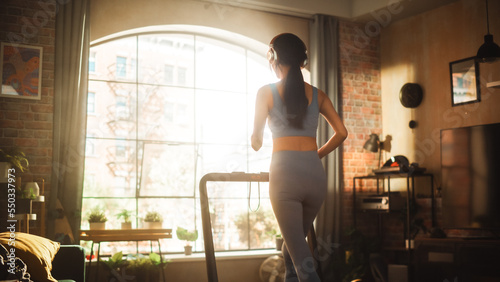 Beautiful Athletic Sports Woman Running on a Treadmill at Her Home Gym. Energetic Workout Female Training while Listening Podcast or Music in Headphones. Apartment with Window View. Side Back View © Gorodenkoff