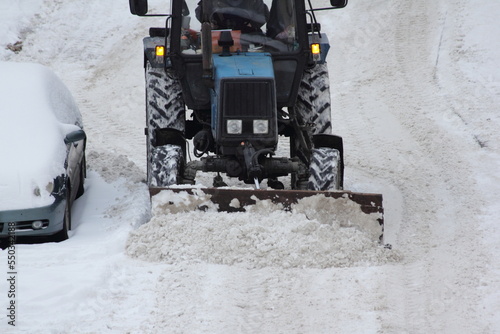 Wheeled bulldozer tractor removes a snow with scraper shovel blade snowplow on road near parked car. parked vehicles interfere with snow removal after heavy snowfall at winter. Front top view