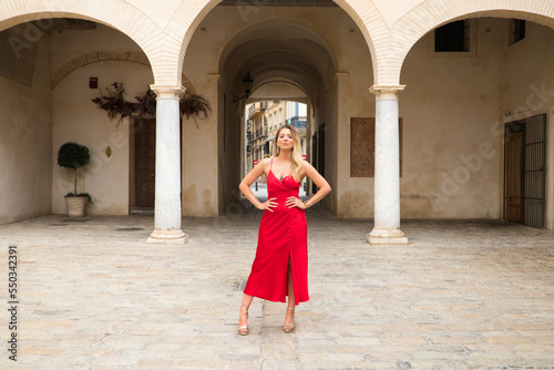 Young, blonde, beautiful woman in a red dress is visiting seville. The woman poses for the camera very elegant and like a model in the typical streets of the city. Holidays and travels