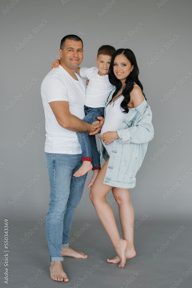 Pregnant couple, a young family with a young son, stand on a gray background, laughing and waiting for their unborn child Pregnant couple, a young family with a young son, stand on a gray background, laughing and waiting for their unborn child