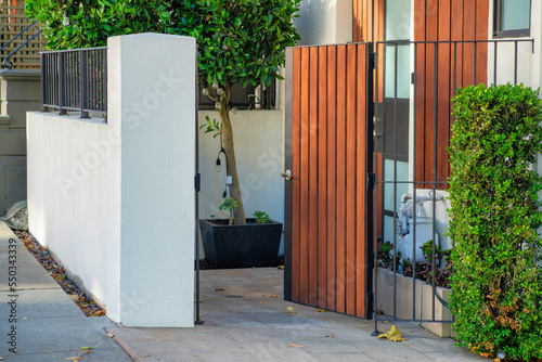 Open courtyard gate with vertical wooden pannels on frence with cement white wall and front yard trees in a neighborhood downtown