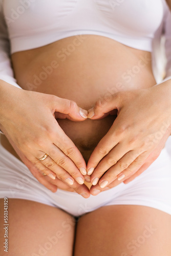 Woman with a big bare tummy is sitting on a bed in her underwear with a bare stomach. A pregnant lady and her husband hold their palms in the shape of a heart on their stomach.