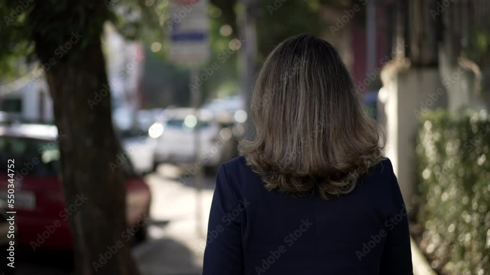 Back of person walking in urban street sidewalk. One Woman walks in city during bright daylight Back of person walking in urban street sidewalk. One Woman walks in city during bright daylight