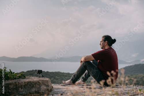 A young man peacefully meets the sunset in a garden with a view of the mountains and the sea.