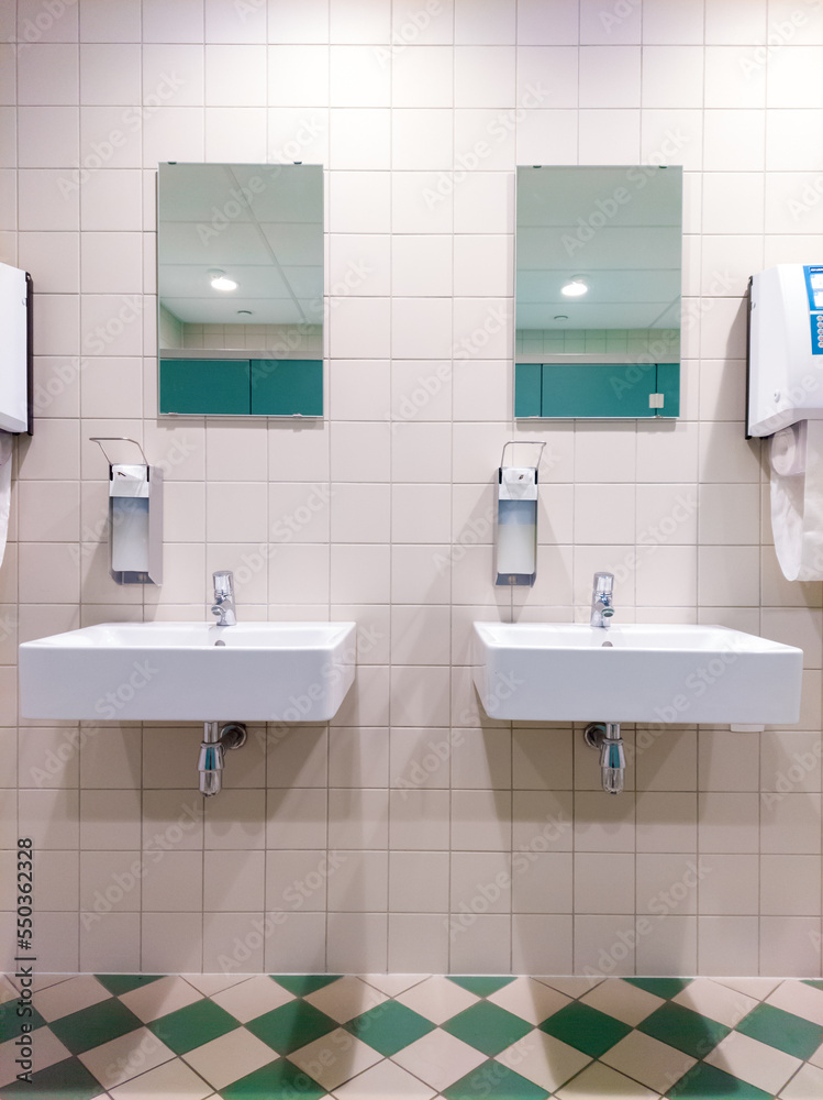 Twin sinks in sanitairy facility with mirrors, soap dispensers and towels. Nice shading pattern created by ceiling lighting. Image taken indoors in Leuven, Belgium. Twin sinks in sanitairy facility with mirrors, soap dispensers and towels. Nice shading pattern created by ceiling lighting. Image taken indoors in Leuven, Belgium.