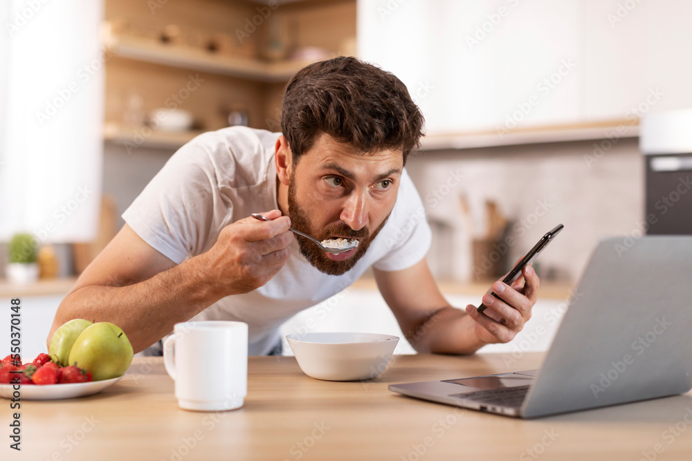 Busy serious adult caucasian man with beard in t-shirt eats porridge, uses smartphone, watches video on laptop Busy serious adult caucasian man with beard in t-shirt eats porridge, uses smartphone, watches video on laptop