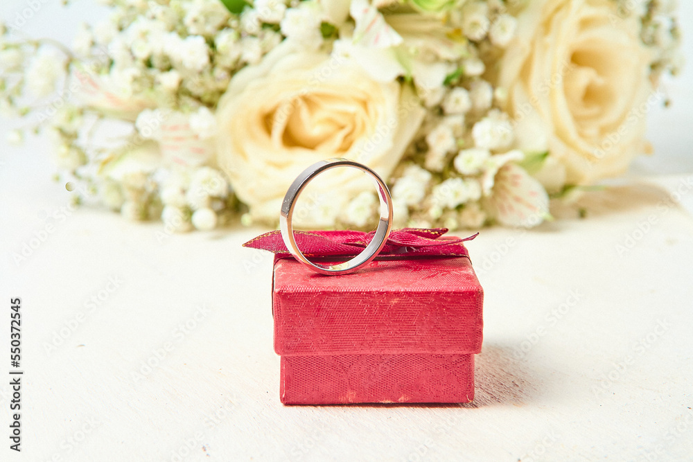 A white gold ring stands on a red gift box and a bouquet of flowers on a white textured tree background, holiday A white gold ring stands on a red gift box and a bouquet of flowers on a white textured tree background, holiday