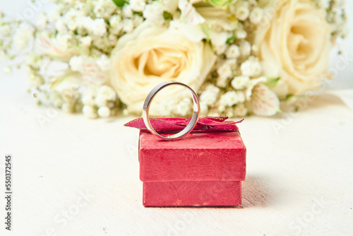 A white gold ring stands on a red gift box and a bouquet of flowers on a white textured tree background  holiday