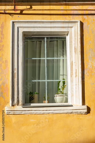 Part of the decoration of the facade of the building and the material from which it is built; a yellow wall with an old window in which there is a flower pot