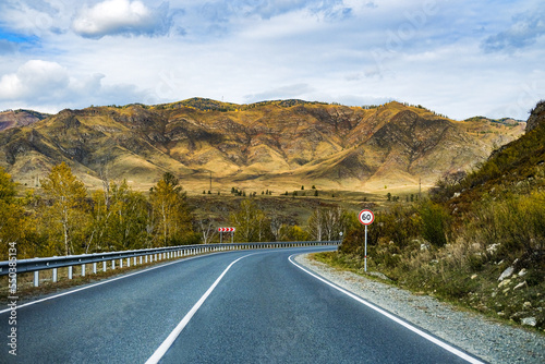 Serpentine asphalt road among high snow-capped mountain peaks, yellow desert, autumn green forest and blue sky. Car on the highway against the background of a mount landscape.