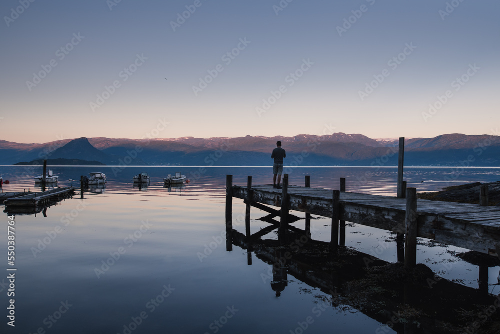 A young man standing on a wooden pier at sunset watching the boats swaying on a Norwegian fjord. Norwegian landscape with lake and mountain. Travel landscape. Norway, Hardangerfjordvegen