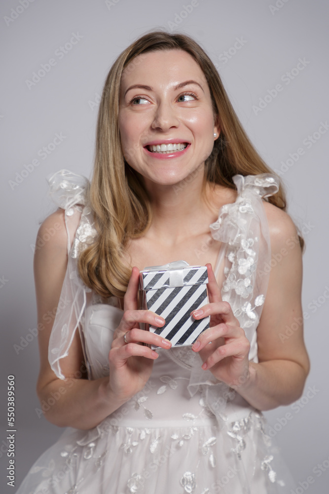 White european young woman with blonde hair in a white dress opens a gift and looks at the gift with a happy smile isolated on gray background White european young woman with blonde hair in a white dress opens a gift and looks at the gift with a happy smile isolated on gray background