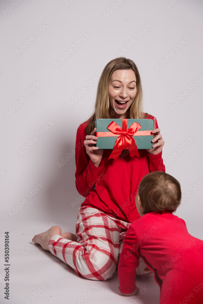 White european young woman with blonde hair in red New Year's pajamas with a gift in her hands smiles happily and looks at the child who is crawling towards her isolated on gray background White european young woman with blonde hair in red New Year's pajamas with a gift in her hands smiles happily and looks at the child who is crawling towards her isolated on gray background