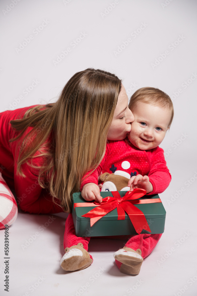 White european young woman with blonde hair with a small child in red New Year's pajamas, mother kisses the child on the cheek isolated on gray background White european young woman with blonde hair with a small child in red New Year's pajamas, mother kisses the child on the cheek isolated on gray background