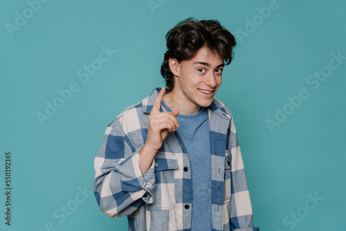 Teenager boy wearing blue plaid shirt over isolated background rises index finger, pensive expression. Smiling with cheerful face. Idea concept. Clever teen isolated on turquoise studio backdrop..