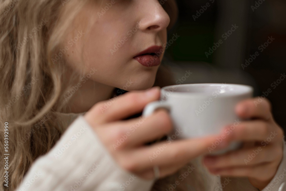 White blonde girl in a warm sweater drinks tea in a coffee shop. In a cafe, coffee is poured into a white mug. Hot tea for morning breakfast. Warming coffee in the restaurant on a winter evening. High White blonde girl in a warm sweater drinks tea in a coffee shop. In a cafe, coffee is poured into a white mug. Hot tea for morning breakfast. Warming coffee in the restaurant on a winter evening. High