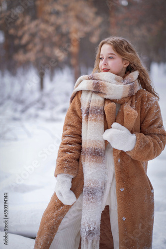 A white blonde girl in a knitted hat, a long scarf and a fur coat walks through the forest and smiles. Woman catches snow in white mittens. Winter is on the street. High quality photo. Christmas Eve