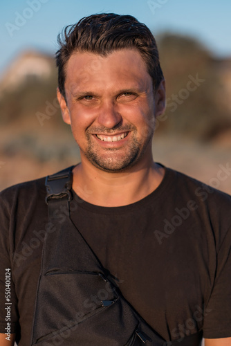 A handsome brunette man in a black t-shirt smiles and looks at the camera against the backdrop of mountains and a blue sky. High quality photo.