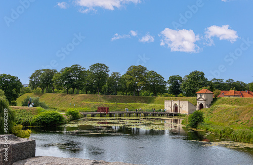 Copenhagen, Denmark - July 24, 2022: Landscape, Green dirt ramparts and moat east of south entrance with bridge into historic military base Kastellet under blue sky. Red roofs