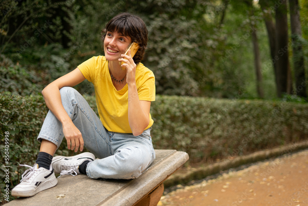 young beautiful hispanic woman talking on her phone and smiling sitting on a bench in a park