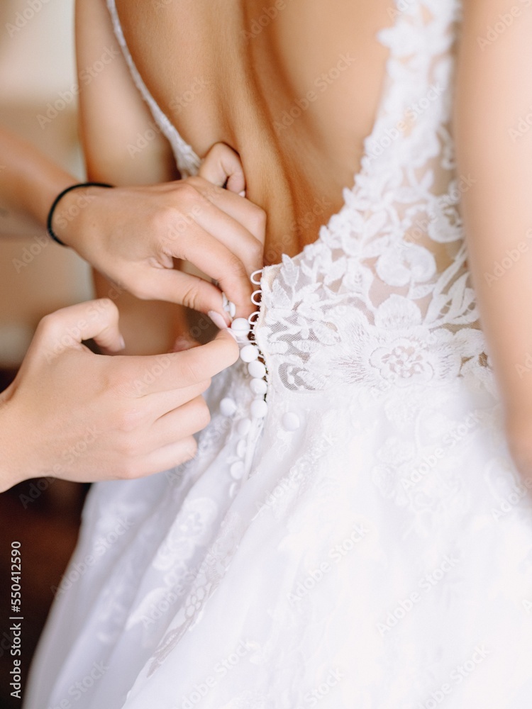 Close up hands of bridesmaids fastening the buttons on wedding dress of bride before wedding ceremony