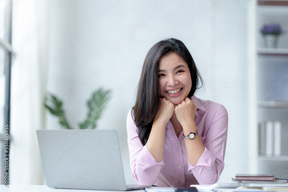 Smiling beautiful young Asian businesswoman sitting with laptop and computer while doing some paperwork at the office. Looking at camera. Smiling beautiful young Asian businesswoman sitting with laptop and computer while doing some paperwork at the office. Looking at camera.