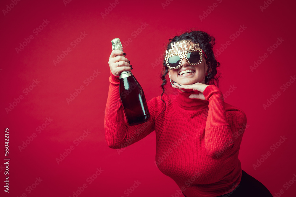 Excited girl smiles having fun with happy new year black and gold glasses, sparkiling champagne bottle, red background, sweater and black skirt Excited girl smiles having fun with happy new year black and gold glasses, sparkiling champagne bottle, red background, sweater and black skirt