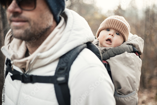 Sporty father carrying his infant son wearing winter jumpsuit and cap in backpack carrier hiking in autumn forest