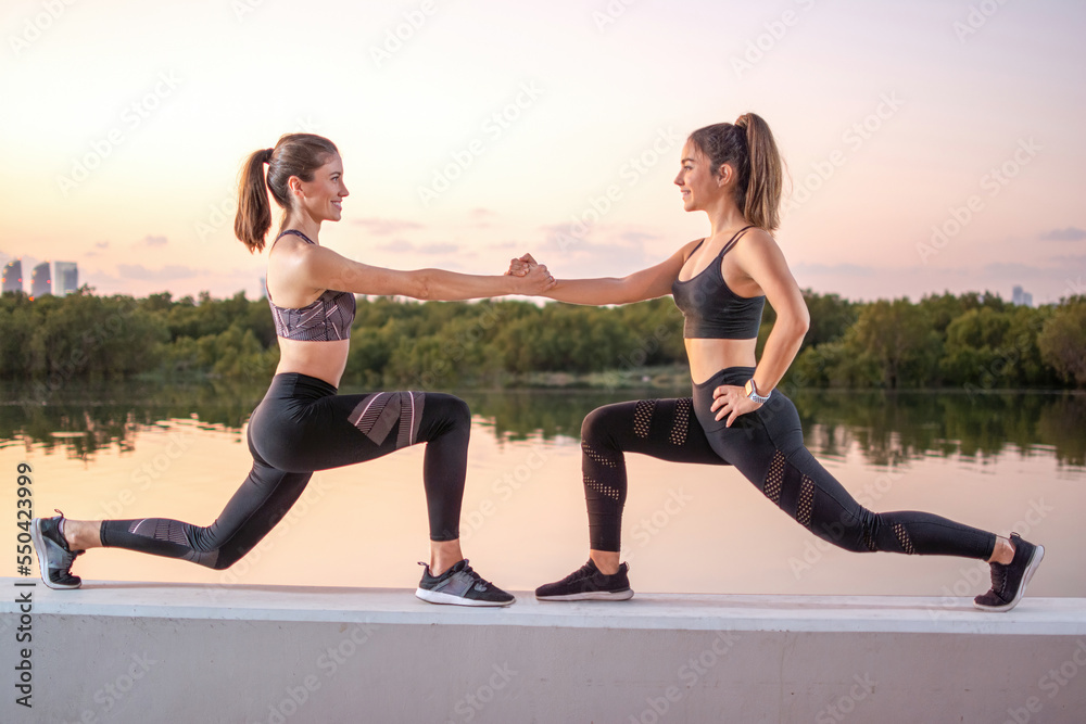 Two attractive young women holding hands while balancing in lunge position outdoors.
