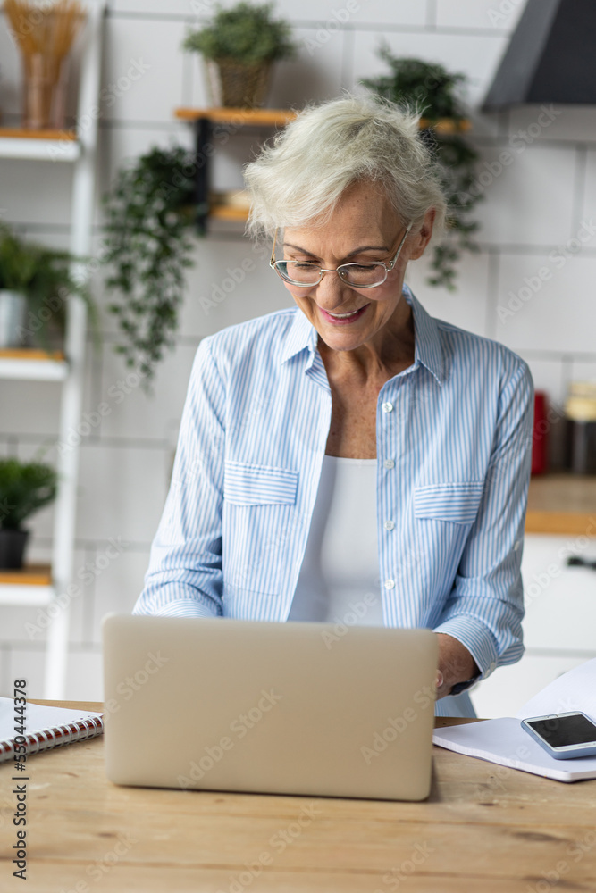 Attractive senior woman working at home in the kitchen using her laptop drinking tea. Concept of mature female using internet and technology for learning, shopping, bank services, freelance job Attractive senior woman working at home in the kitchen using her laptop drinking tea. Concept of mature female using internet and technology for learning, shopping, bank services, freelance job