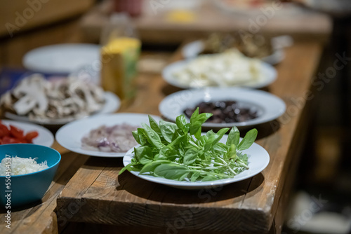 Ingredients for a home made pizza, such as greenery, ham, cheese, mushrooms, prepared on plates.