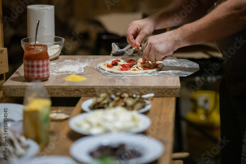 Man preparing a delicious fresh home made pizza. Person topping pizza dough with mushrooms, tomatoes, cheese and so on.