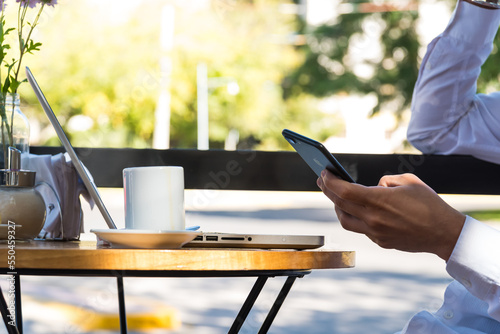 Man looking at his cell phone while sitting outdoors, laptop and coffee on the table