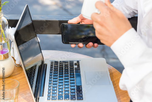 person having coffee while looking at their smartphone, laptop on the table