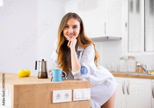 Cheerful woman in man's shirt drinking fresh coffee in kitchen at home.