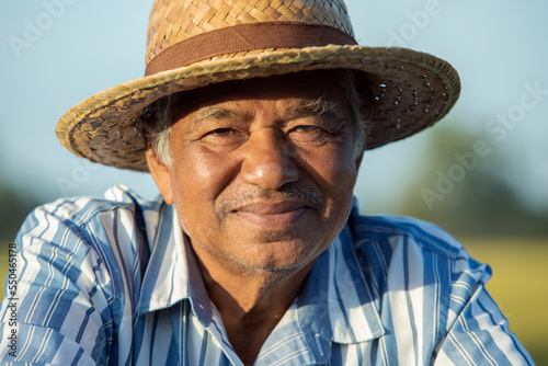 Close-up of elderly Asian farmer wearing a shirt and hat looking at the camera sitting alone outdoors at spring. Portrait farmer, Thailand © Pituk