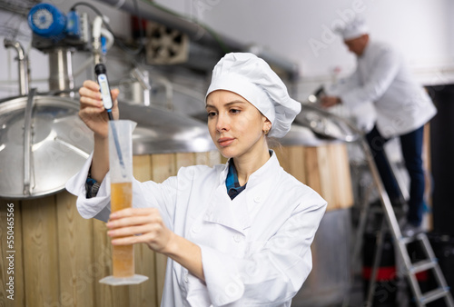 Focused skilled young female brewery worker using digital hydrometer for measuring craft beer density..