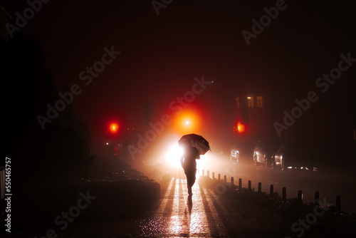 A man with an umbrella walks through the night foggy city. The rays of car headlights and traffic lights illuminate the silhouette of a person. Soft focus.