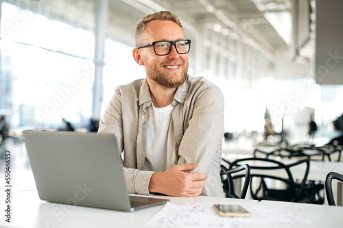 Positive smart successful caucasian man, with glasses, in stylish casual wear, IT specialist, company seo, freelancer, sits at a desk with a laptop in coworking, looks away, smiles happily