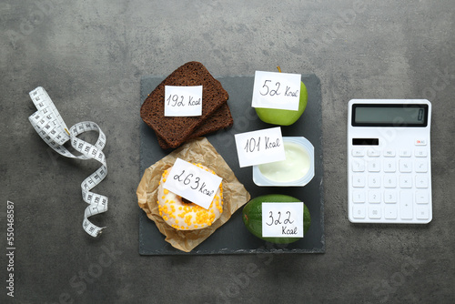 Calculator and food products with calorific value tags on dark grey table, flat lay. Weight loss concept