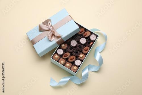 Open box of delicious chocolate candies and light blue ribbon on beige background, flat lay