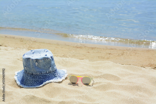 Fototapeta Naklejka Na Ścianę i Meble -  Jeans hat and sunglasses on sand near sea, space for text. Beach accessories