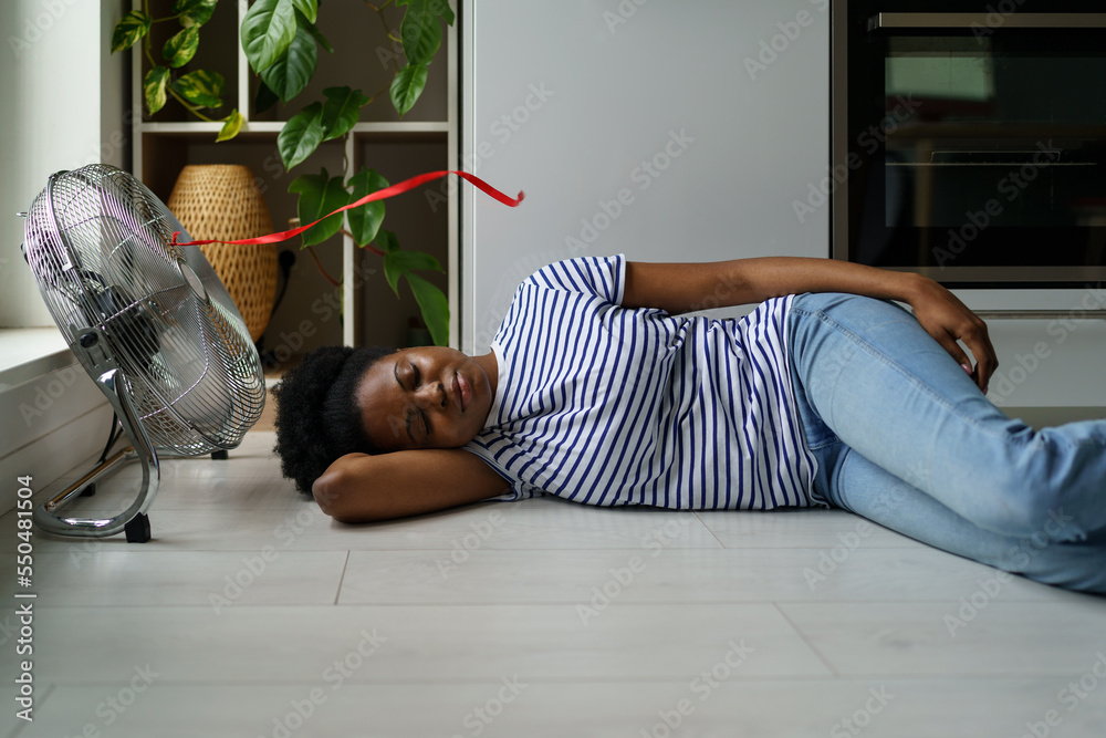Overheated African woman lying on floor sleeping with electric fan on