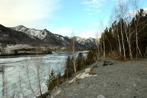 View from a high rocky bank of a frozen river surrounded by snow-capped mountains.