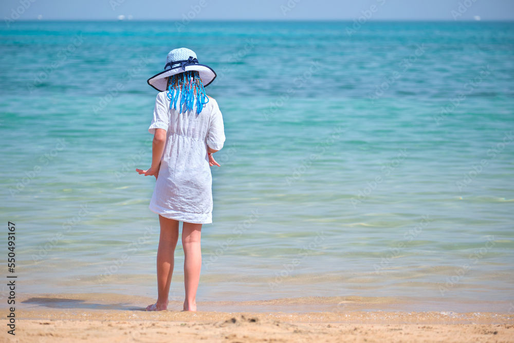 Rear view of child girl in white summer dress and sunshade hat with blue ribbons in long braids standing barefoot on sandy beach enjoying sea view