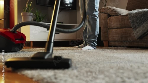 Home cleaning service and chores. Household woman vacuums carpet near sofa in apartment. Low angle view of vacuum cleaner and female legs. Tracking left shot. photo