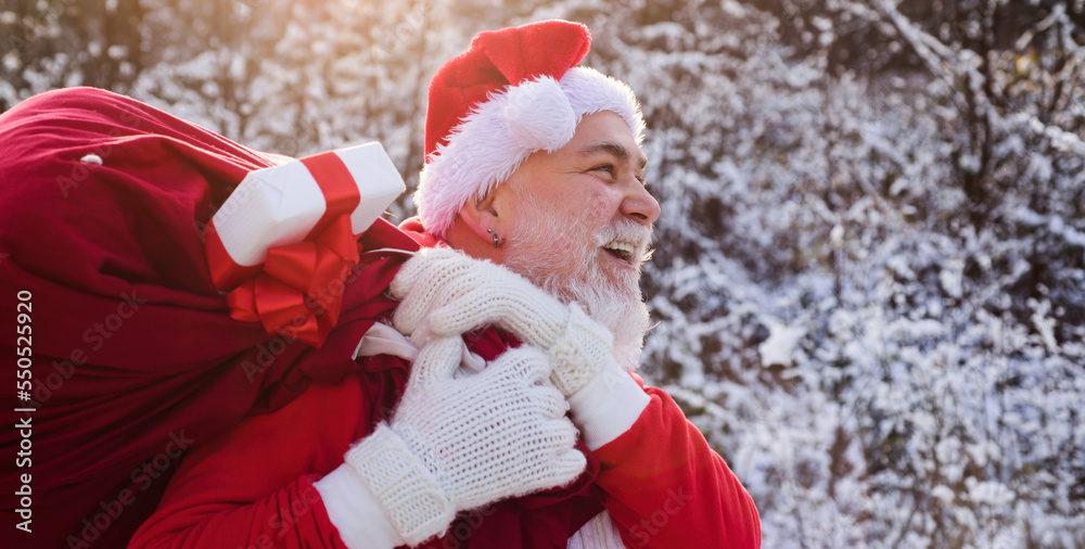 Obraz premium Santa claus with sack full of christmas presents outdoor. Santa Claus walk in the winter snow park.
