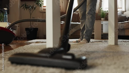 Home cleaning service and household chores. Housewife woman vacuums carpet under table in apartment. Close-up of vacuum cleaner and female legs, low angle view. Tracking left shot.