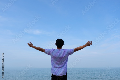 man on beach hold hands arms up, rear view guy wearing summer hat, standing back looking to sea blue sky horizon, vacation concept of freedom travel ocean