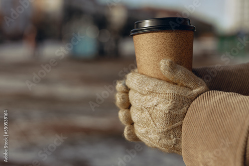 Crop woman in knitted gloves holding paper cup of takeaway coffee and warming hands on sunny day in winter in city
